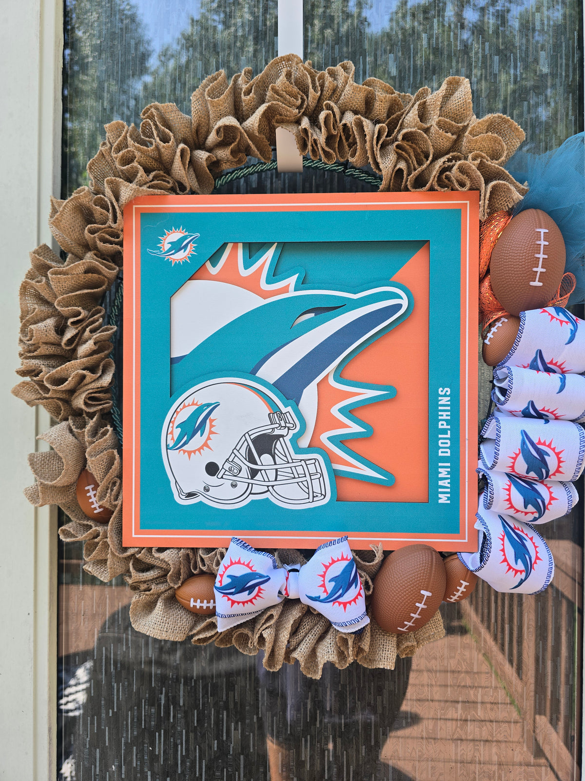 A Miami Dolphins themed decorative wreath hanging on a front door. The wreath features a large brown burlap ruffle border, a central square orange and teal plaque with a dolphin logo and helmet, brown football-shaped ornaments, and white bows printed with the Miami Dolphins logo.