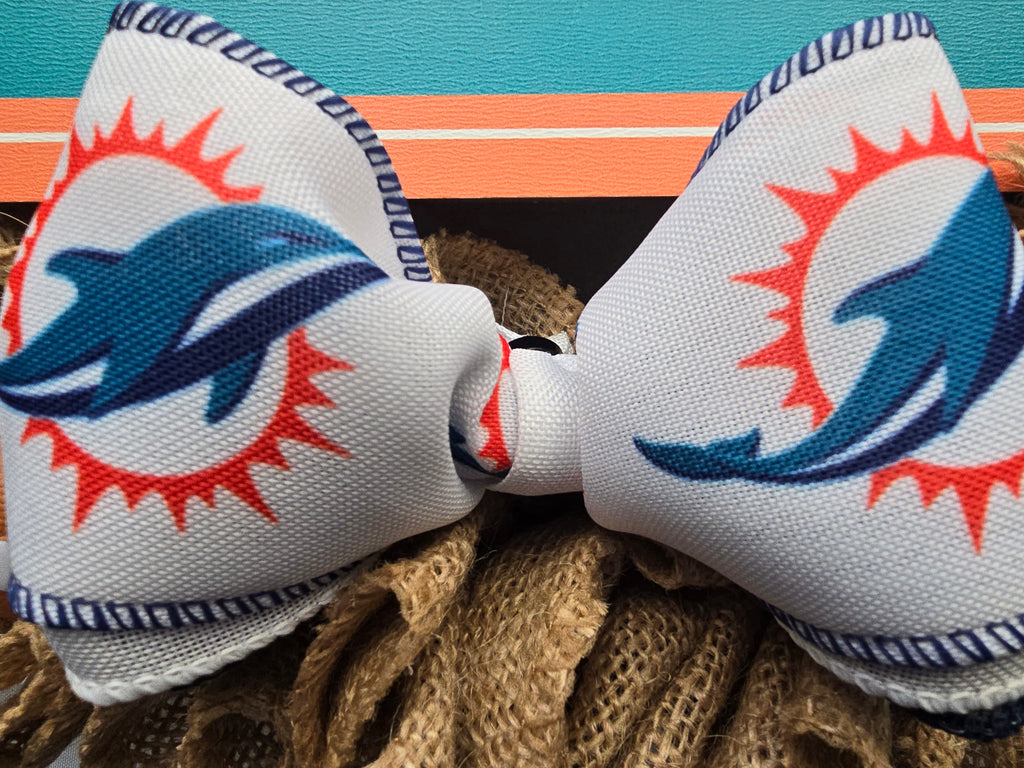 A white fabric bow featuring the Miami Dolphins logo, styled on a burlap wreath against a teal and orange background.